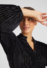 Blusa nera con trama, strisce verticali, colletto e chiusura con bottoni. Maniche lunghe a sbuffo con polsini elasticizzati.