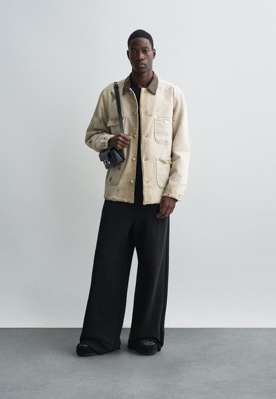 Veste en denim beige avec un col et des poches marron, portée sur une chemise noire. Pantalons noirs larges et petit sac bandoulière noir.