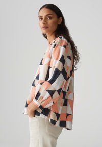 Camicia multicolore con un motivo geometrico, caratterizzata da tonalità di arancione, blu navy e beige. Vestibilità ampia con maniche a tre quarti e scollatura rotonda.