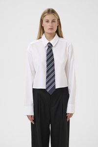 Mlada ženska z dolgimi blond lasmi, oblečena v belo kratko majico, črtast temno modri neč, in hlače z visokim pasom v črni barvi, ki stoji ob beli ozadju.