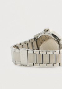 Ασημένιο μεταλλικό βραχιόλι ρολογιού με κούμπωμα και στρογγυλή κάσα ρολογιού που βρίσκεται σε λευκή επιφάνεια, δείχνοντας την πίσω πλευρά.