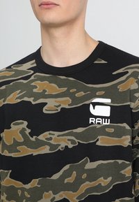 Svart sweatshirt med ett camouflage-mönster i grön och beige. Har en rund halsringning och en vit "RAW"-logotyp på bröstet.