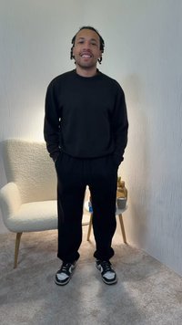 Ensemble sweat-shirt et pantalon de survêtement noir, assorti à des baskets noires et blanches. Debout sur un tapis près d'un fauteuil texturé de couleur claire.