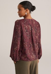 Femme aux cheveux bruns en chignon portant des boucles d'oreilles en croissant argentées, un chemisier à motifs marron à manches longues et un pantalon vert olive, vue de dos.