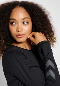 Czarna koszula z długim rękawem, o gładkiej fakturze i zaokrąglonym dekolcie. Z lewej strony rękawa szare akcenty w kształcie szewronu.