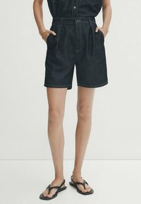 Shorts en denim bleu foncé, taille haute avec des plis sur le devant. Fini avec des coutures contrastées et une texture lisse. Port de sandales noires.