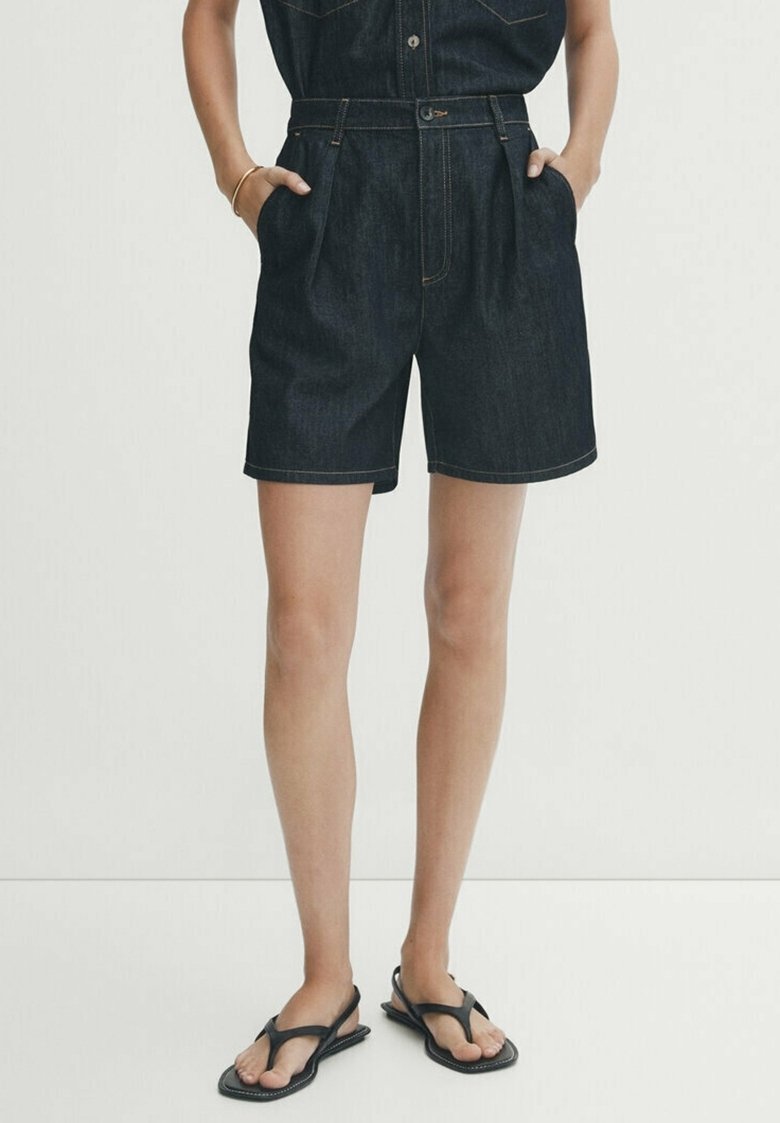 Shorts en denim bleu foncé, taille haute avec des plis sur le devant. Fini avec des coutures contrastées et une texture lisse. Port de sandales noires.