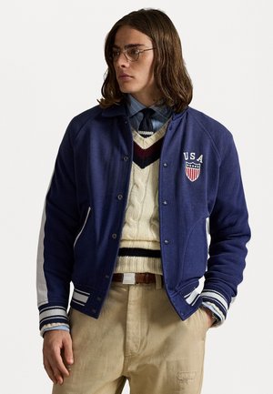 Dark blue varsity jacket with white accents, featuring a USA patch. Worn over a beige knit sweater and collared shirt, paired with khaki pants.