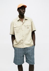 Άνδρας με μπεζ πουκάμισο με κοντά μανίκια και κουμπιά, πορτοκαλί καπέλο και μπλε καρό σορτς, στέκεται με τα χέρια στις τσέπες μπροστά σε λευκό φόντο.