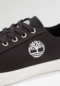 Baskets en toile noire avec lacets et œillets blancs, arborant un logo brodé d'un arbre et des lignes horizontales sur le côté. Semelle blanche.