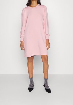 Kvinde iført en pastel pink minikjole med lange ærmer og puffede skuldre, kombineret med grå glitrende sokker og sorte spidse sko.