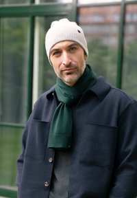Giacca in misto lana blu navy, maglione grigio sotto, sciarpa in maglia verde, berretto grigio chiaro e una pelle liscia con barba corta.