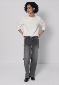 Vitsvart sweatshirt med långa ärmar, kombinerad med grå vida jeans. Svarta skor, stående position, neutral bakgrund.