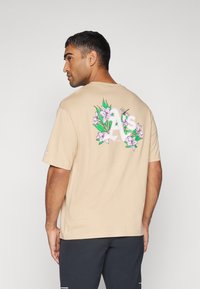 Camiseta de algodón beige con diseño floral y gráfico de "A's" en la parte posterior, con mangas cortas y un corte relajado.