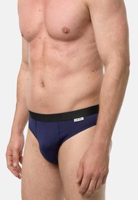 Męski tors w granatowych bokserkach z czarnym paskiem w talii i małą metką, stojący na jednolitym jasnym tle.