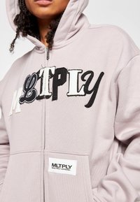 Helle rosa Zip-Hoodie mit großem, gesticktem "MLTPLY"-Text in Schwarz und Weiß. Verfügt über eine Kapuze und ein Markenetikett am Saum.