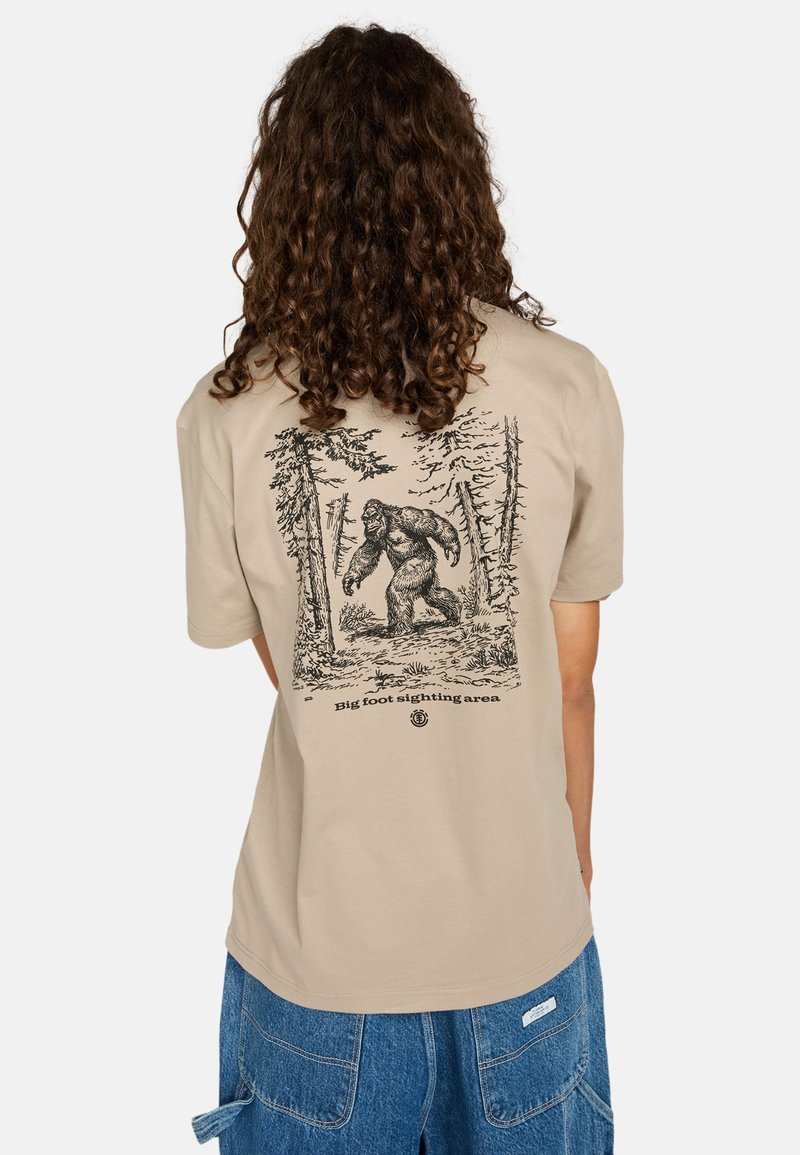 Camiseta de algodón beige con una ilustración impresa en negro de Bigfoot entre árboles en la parte posterior. Mangas cortas, corte relajado.