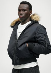 Μαύρο μπουφάν τύπου bomber με κουκούλα με γούνα, φερμουάρ μπροστά, πλέκι μανίκια και πλαϊνές τσέπες. Φοριέται πάνω από ένα λευκό T-shirt.