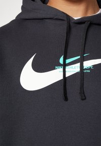 Haut à capuche noir en tissu doux, arborant un grand logo Nike blanc et un texte turquoise plus petit. Il possède une capuche avec cordon de serrage et une poche frontale.