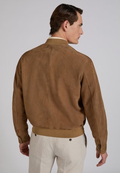 Man draagt een bruine suède bomberjack en lichtbeige broek, staat met zijn rug naar de camera tegen een effen achtergrond.