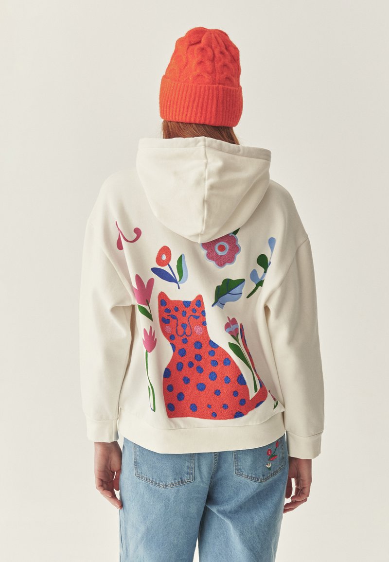 Sudadera crema con un gran gráfico de un gato colorido que tiene puntos azules y diseños florales. Combinada con jeans de un azul claro y un gorro de punto naranja.