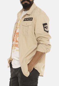 Chemise khaki à boutons avec deux poches poitrine, arborant un patch noir marqué "VON DUTCH" et un patch "ARMY" sur la manche. La couche intérieure est un t-shirt graphique blanc.