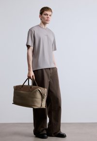 Szürke póló, bő barna nadrág, és egy nagy bézs színű sporttáska barna díszítéssel. A táskán két fogantyú és cipzáras záródás található.