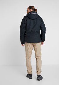 Zwarte Columbia-jas met capuchon, beschikt over verstelbare manchet en een logo op de achterkant. Gepaard met beige broeken en zwarte schoenen.