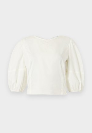Blouse blanche à manches longues avec manches bouffantes et poignets élastiques, présentée sur un fond gris clair uni.
