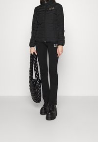 Czarna pikowana kurtka z teksturowanym wzorem, zapinana na zamek, z logo. Dopasowane czarne legginsy i pleciona torba na ramię, uzupełnione butami na platformie.