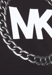 Gros plan d'un collier en chaîne argenté posé sur un tissu noir avec de grandes lettres blanches "MK" imprimées dessus.
