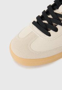 Beige sneaker with a textured suede and leather upper, black laces, and a gum rubber sole. Rounded toe and stitched detailing.
