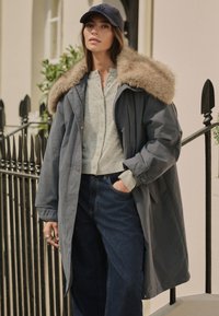 Parka grise avec col en fourrure, associée à une chemise gris clair à motifs et un jean bleu foncé. Devant une clôture en fer forgé.