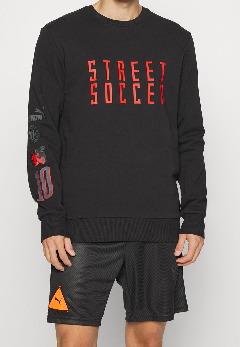 Sort sweatshirt med "STREET SOCCER" i røde bogstaver. Har grafik på ærmerne og en ribbet halsudskæring. Kombineret med sorte teksturerede shorts.