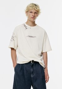 Tricou din bumbac crème cu un design abstract de stropi negri, mâneci scurte și decolteu rotund. Asortat cu pantaloni plisati de culoare bleumarin.