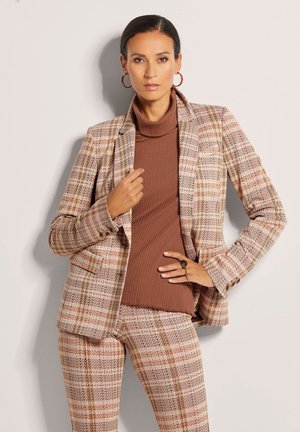 Geruite blazer in roze, bruin en crèmetinten met een getailleerde pasvorm, gelaagd over een bruine ribbeltrui met hoge hals. Bevat een decoratieve ring aan de hand.