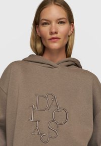 Hoodie in taupe met textuur en verhoogde borduursels met letters in een cirkelpatroon. Zachte stof met een relaxte pasvorm.