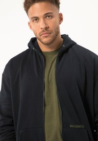 Felpa con zip color navy e cappuccio regolabile, dotata di una maglietta interna verde oliva, dettagli con logo e tasche frontali. Tessuto semplice e texturizzato.