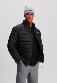 Zwarte gewatteerde jas met een ritssluiting aan de voorkant, hoge kraag en twee zijzakken. Grijze gebreide beanie met het geborduurde logo "KARL LAGERFELD". Grijze spijkerbroek.