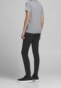 Graues T-Shirt mit kurzen Ärmeln kombiniert mit schwarzen Slim-Fit-Hosen und schwarzen Sneakern mit weißen Sohlen, von hinten betrachtet. Einfaches Design, ohne Muster.