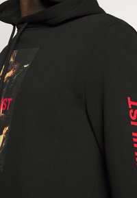 Sudadera negra con capucha que presenta un estampado gráfico rectangular y texto rojo que dice "IST" en la manga izquierda. Tela de algodón suave y texturizada.
