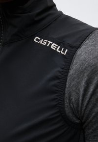 Close-up van een zwarte mouwloos vest met rits en wit "Castelli" logo op de schouder over een grijs shirt met lange mouwen.