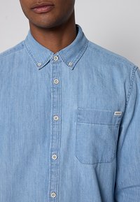 Camicia in chambray azzurro chiaro con colletto button-down, taschino frontale e bottoni a pressione in perlato. Il tessuto ha una texture delicata e un design informale.