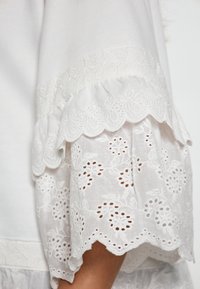 Bluză albă cu dantelă intricate la margine, având margini ondulate și broderie florală. Textură moale și material ușor.