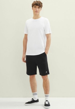 T-shirt en coton blanc, shorts noirs avec poches et baskets noires. Accentué avec des chaussettes rayées blanches et bleues. Tenue décontractée.