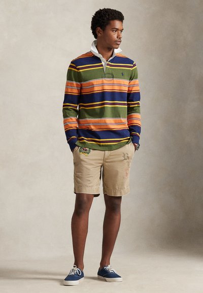 Πολύχρωμο ριγέ πόλο μπλουζάκι σε πράσινο, πορτοκαλί και μπλε με λευκό γιακά. Μπεζ σορτς με κεντημένες λεπτομέρειες. Μπλε αθλητικά παπούτσια.