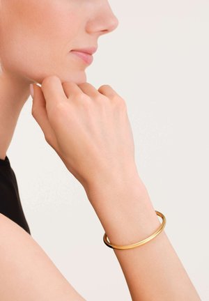 Bracciale in metallo dorato con una superficie liscia e lucida, indossato su una mano che riposa vicino al mento. Il design è circolare e minimalista.