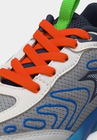 Gros plan sur une basket grise avec des superpositions blanches, des lacets orange vif, des surpiqûres décoratives bleues et une languette en mesh bleu marine avec un tirant vert.