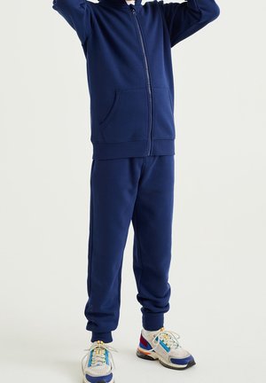 Barn iført en navy blå hættetrøje med lynlås og matchende sweatpants, står med armene løftet, hvide sneakers med blå og røde detaljer.