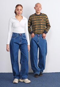 Dos personas están de pie una al lado de la otra. Una lleva una camiseta blanca de manga larga y jeans anchos azules, mientras que la otra lleva un suéter con estampado y jeans similares.